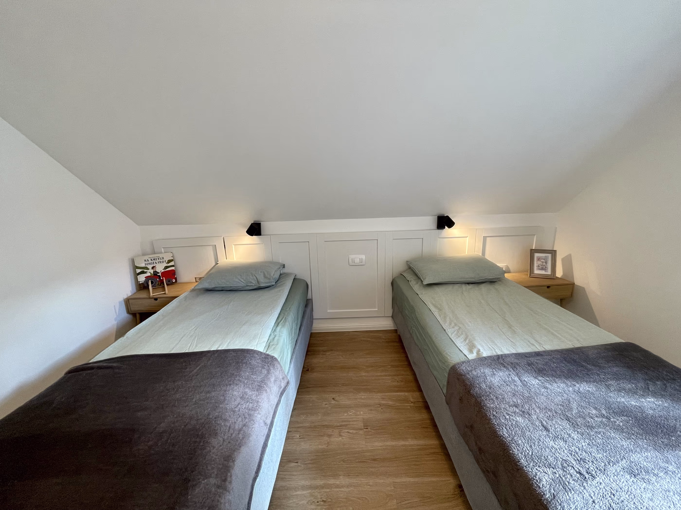 Minimalist bedroom with two single beds with light gray bedding and dark gray throws, each with a wooden nightstand and wall lamps above.