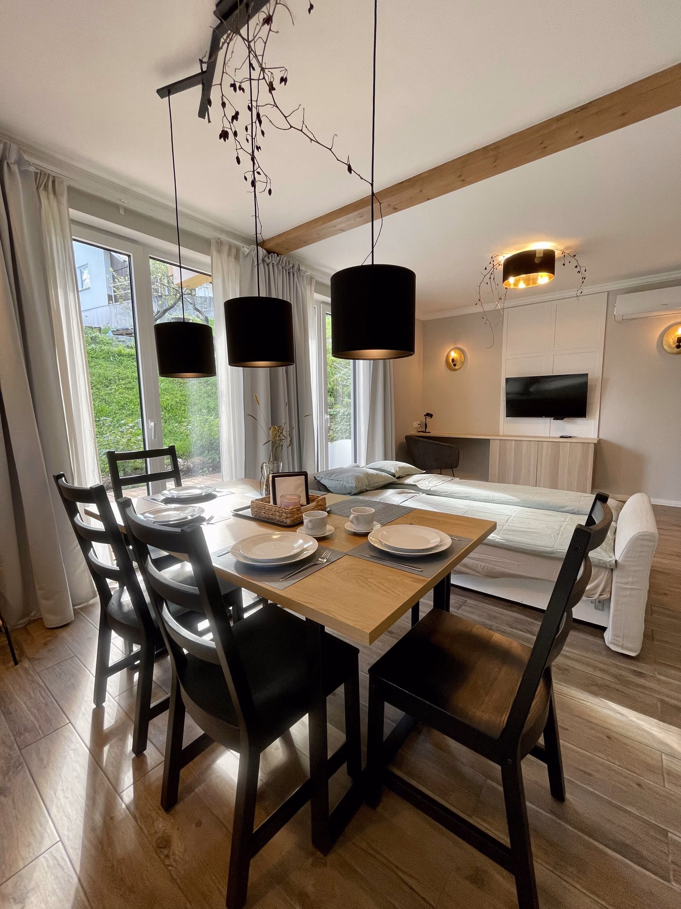 Modern dining area with wooden table set for four, black chairs, pendant lights, and a couch and TV in the background.