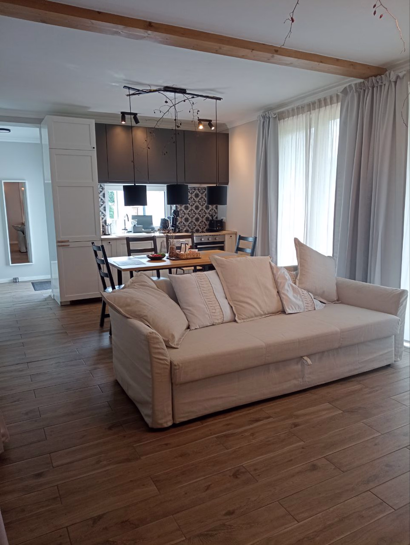 Modern open living space with beige sofa, wooden dining table, black chairs, kitchen with white and dark cabinets, and large curtained window.