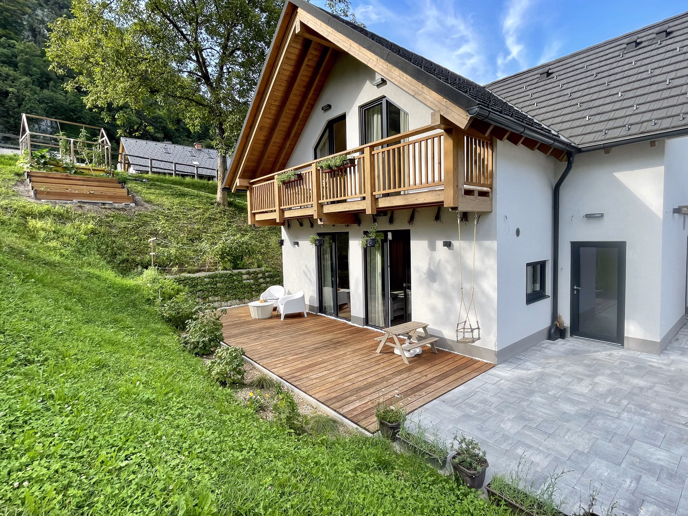 Modern white house with wooden balcony and deck, outdoor seating, a swing, and green lawn on a sunny day.