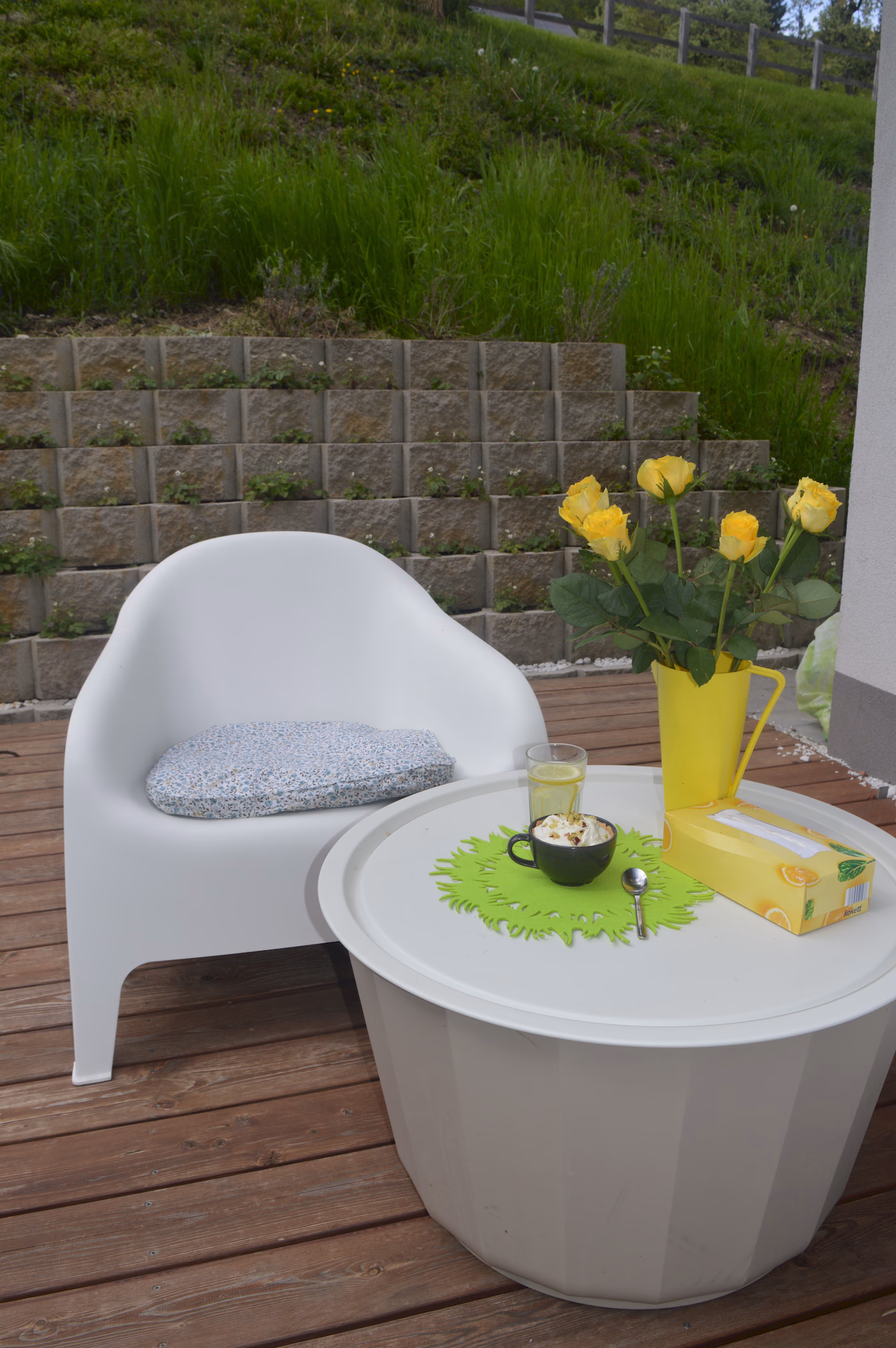 White outdoor chair with a patterned cushion next to a round white table holding a green placemat, a black cup with whipped cream, a glass of lemon water, a yellow vase with yellow roses, and a yellow tissue box on a wooden deck.