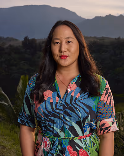 Woman with long dark hair wearing a colorful tropical-patterned dress standing outdoors with mountains in the background at dusk.