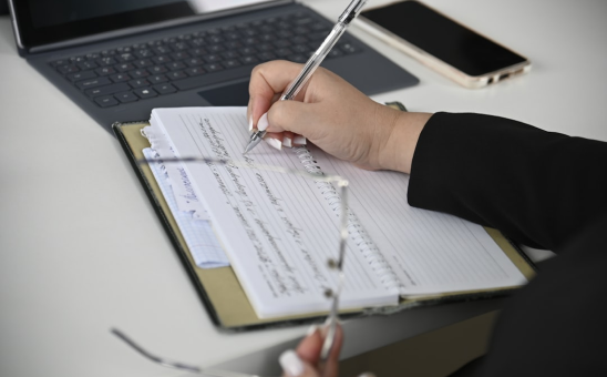 Person writing notes in a notebook with a pen, next to a laptop and smartphone on a white desk.