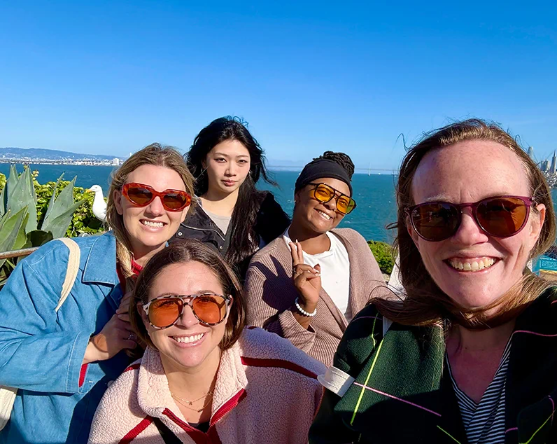 Beyond women wearing sunglasses posing for a selfie by the ocean on a sunny day.
