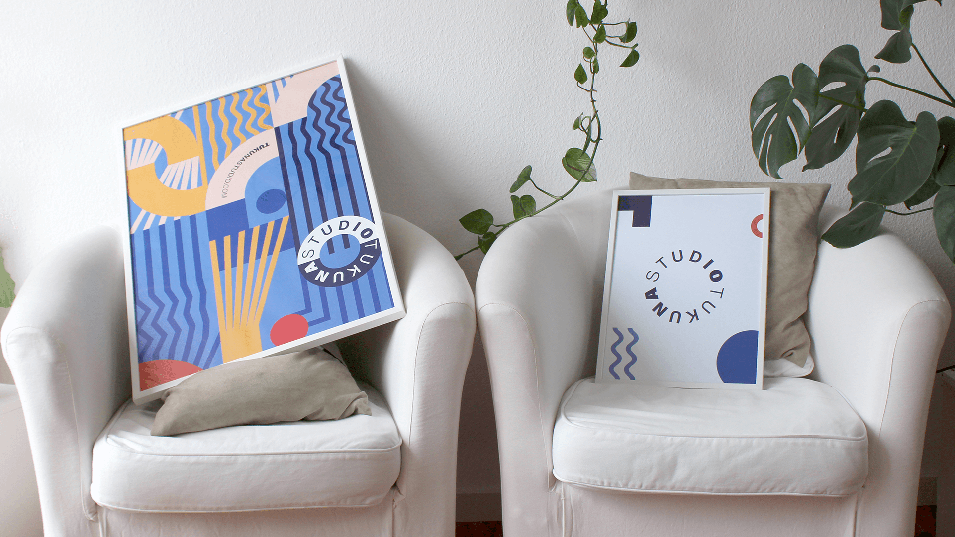 Two framed Tukuna Studio posters displayed on white armchairs beside indoor plants