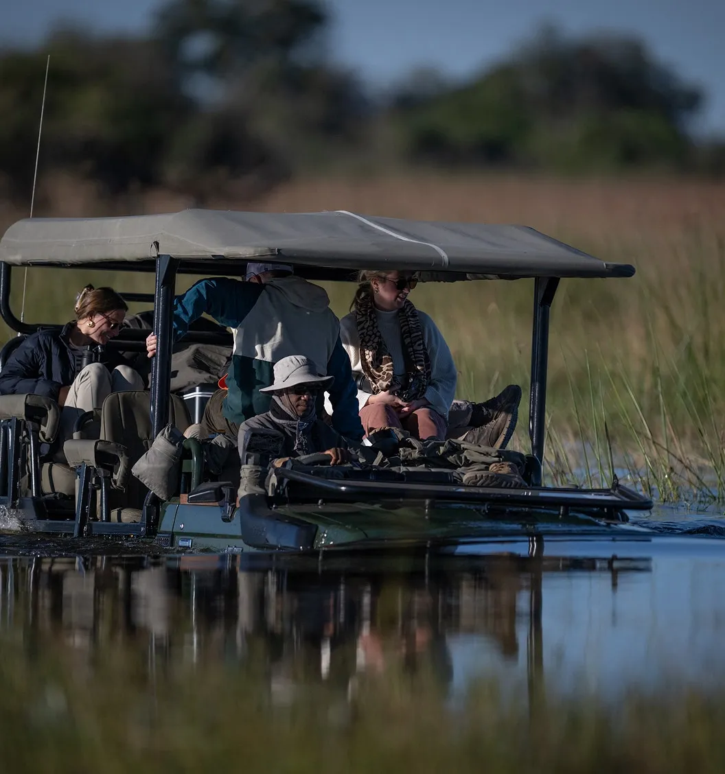 People on a covered safari vehicle driving through shallow water in a natural landscape.