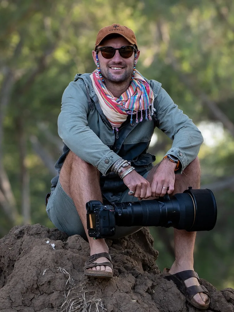Man wearing sunglasses, a cap, and a colorful scarf sitting on a rock outdoors holding a large camera with a telephoto lens.