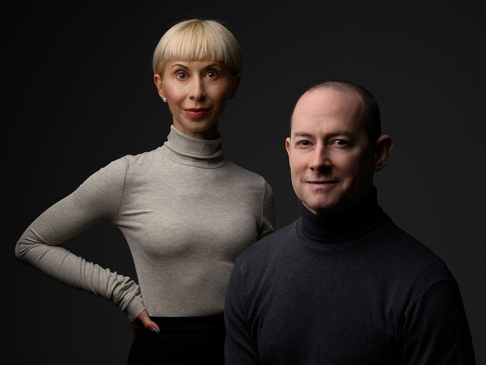 Portrait of a woman with short blonde hair wearing a beige turtleneck standing behind a man with short hair in a dark turtleneck against a dark background.