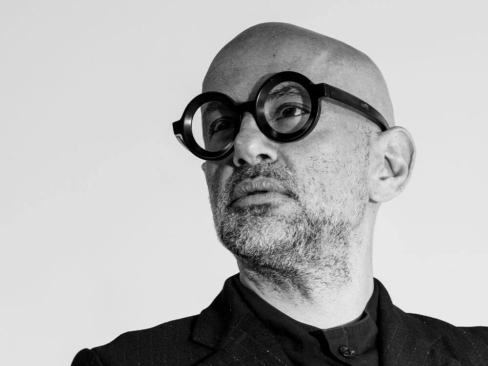 Black and white portrait of a bald man with a short beard wearing round black glasses and a dark suit looking slightly upward.