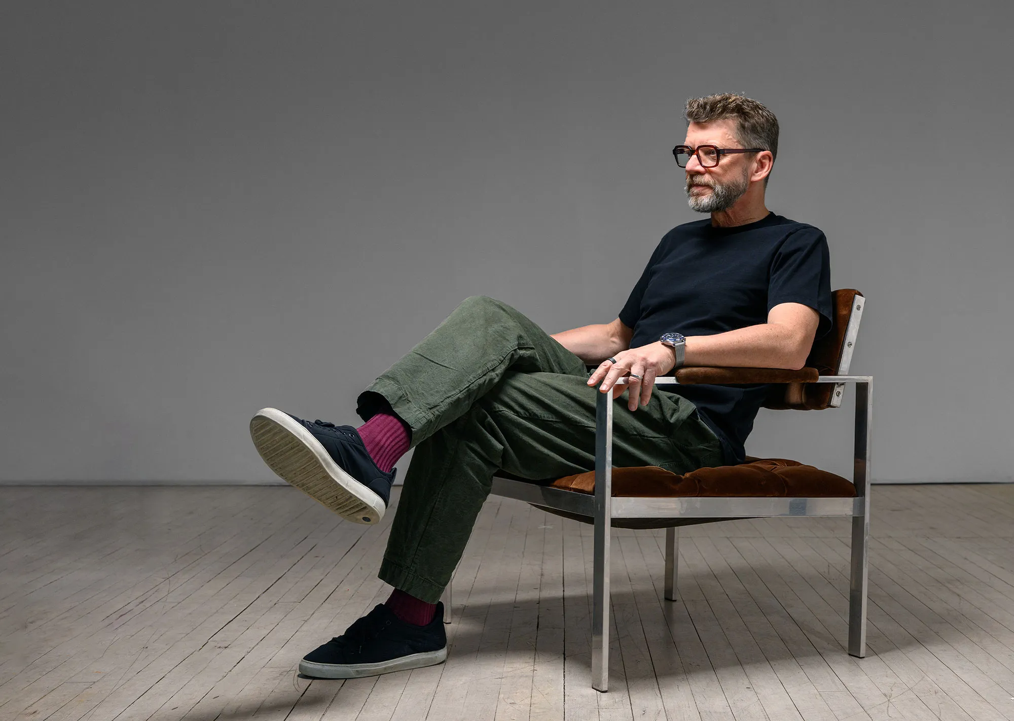 Man with glasses, a black shirt, green pants, and purple socks sitting with legs crossed on a modern metal and brown cushioned chair against a plain gray background.