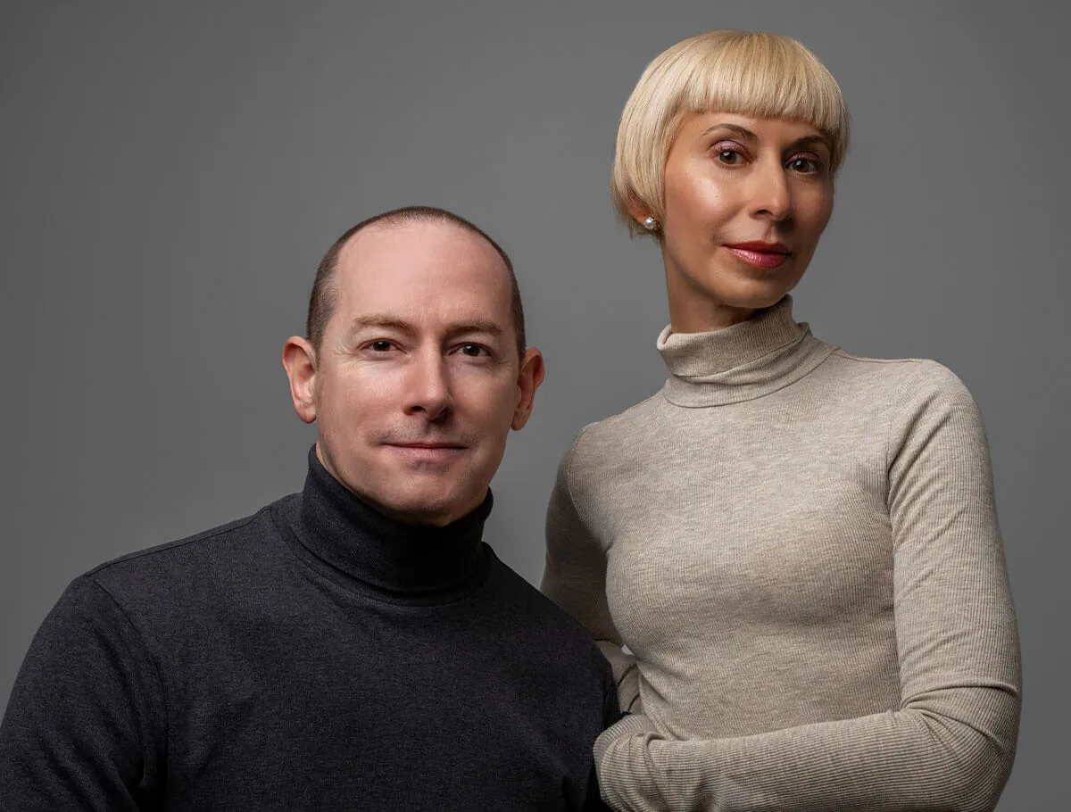 Portrait of a man wearing a black turtleneck and a woman with short blonde hair wearing a beige turtleneck against a gray background.