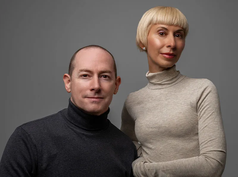Man and woman standing closely together against a gray background, both wearing turtleneck sweaters.