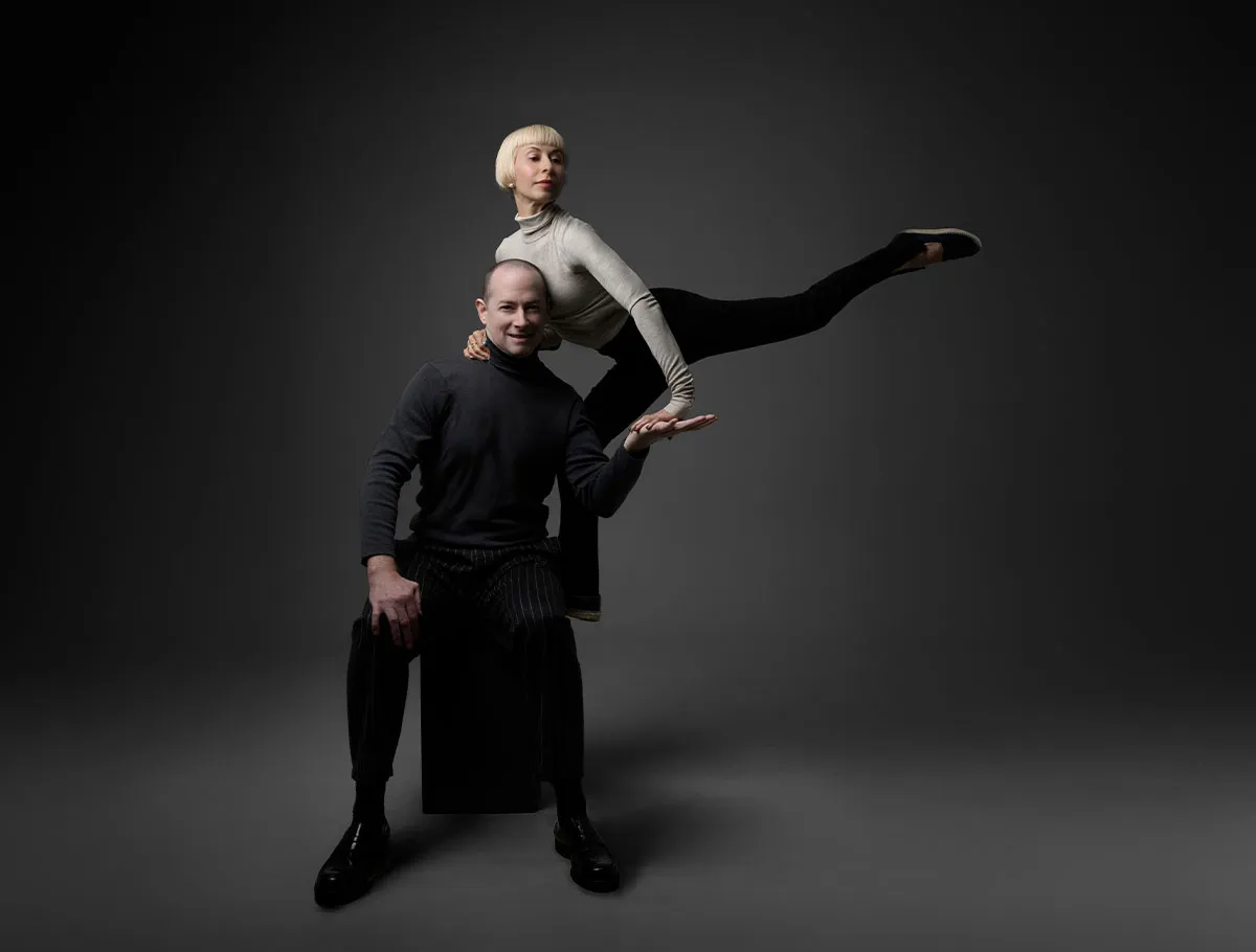 Man seated on a black cube with a woman balancing horizontally on his hand against a dark gray background.