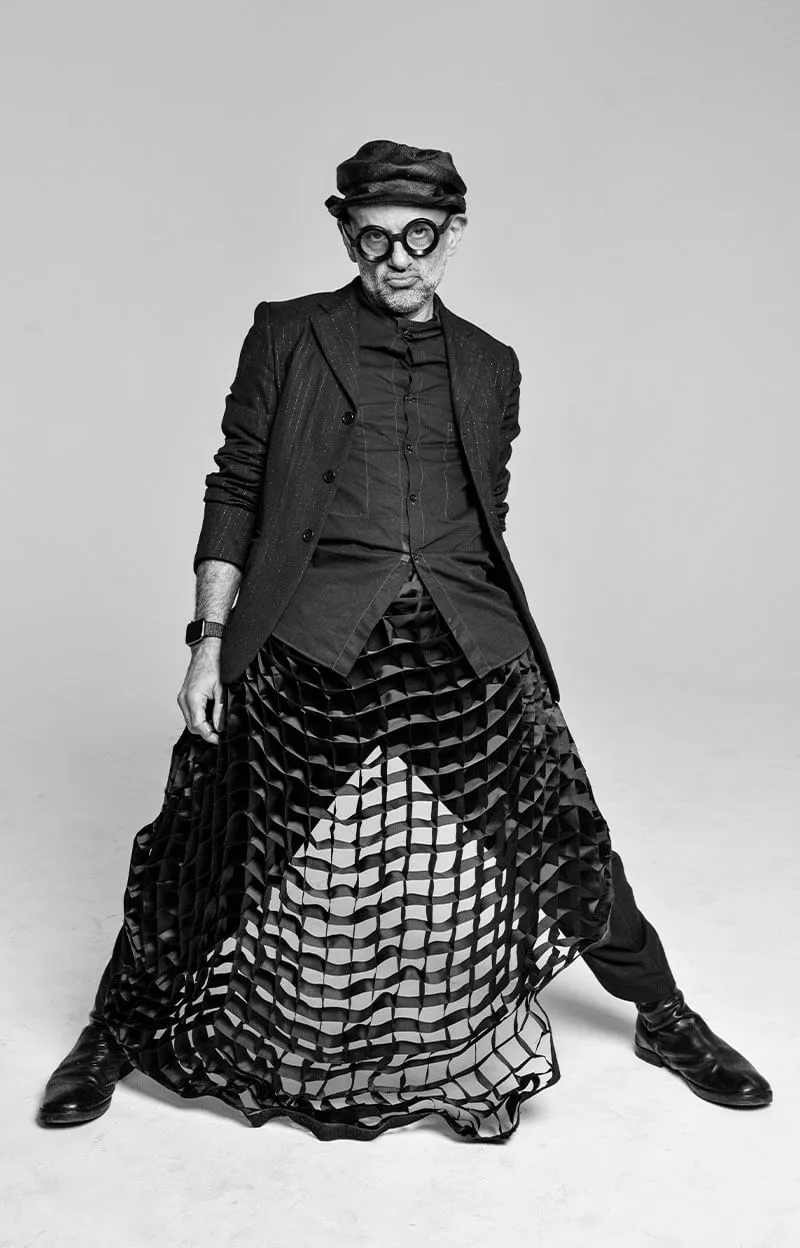 Black and white photo of a man wearing round glasses, a cap, a blazer, and a transparent grid-patterned skirt posing with legs apart.