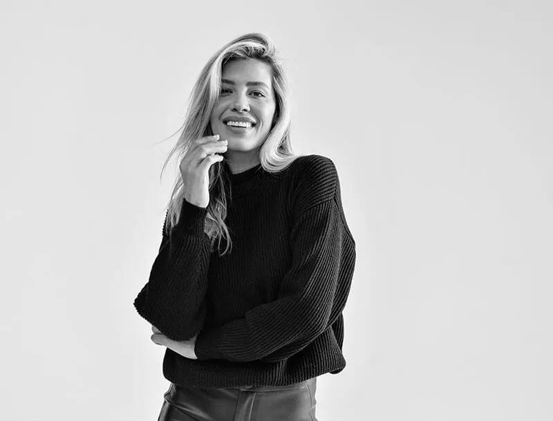 Smiling woman with long hair wearing a black sweater and pants against a plain light background.