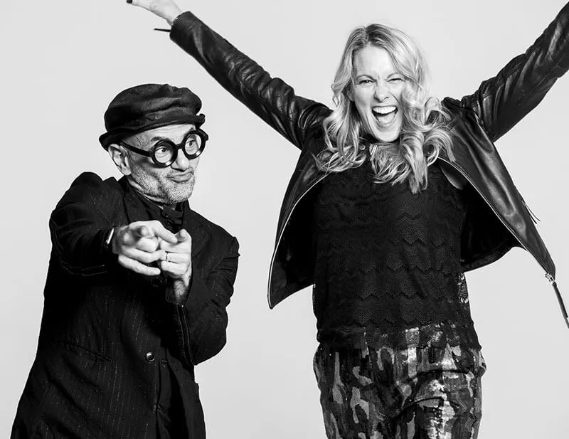 Black and white photo of a man wearing round glasses and a hat pointing at the camera next to an excited woman with raised arms, wearing a leather jacket.
