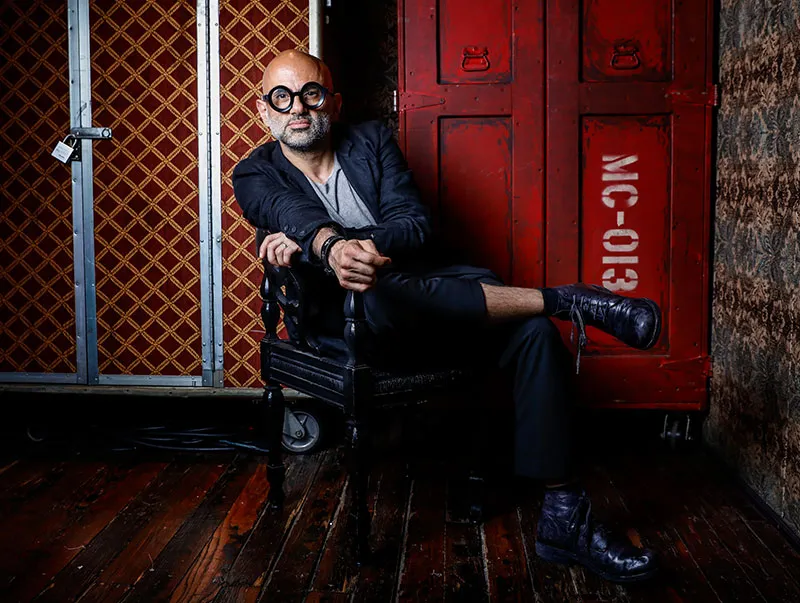 Bald man with a gray beard and round black glasses sitting cross-legged on a dark wooden chair against a backdrop of patterned and red storage cases.