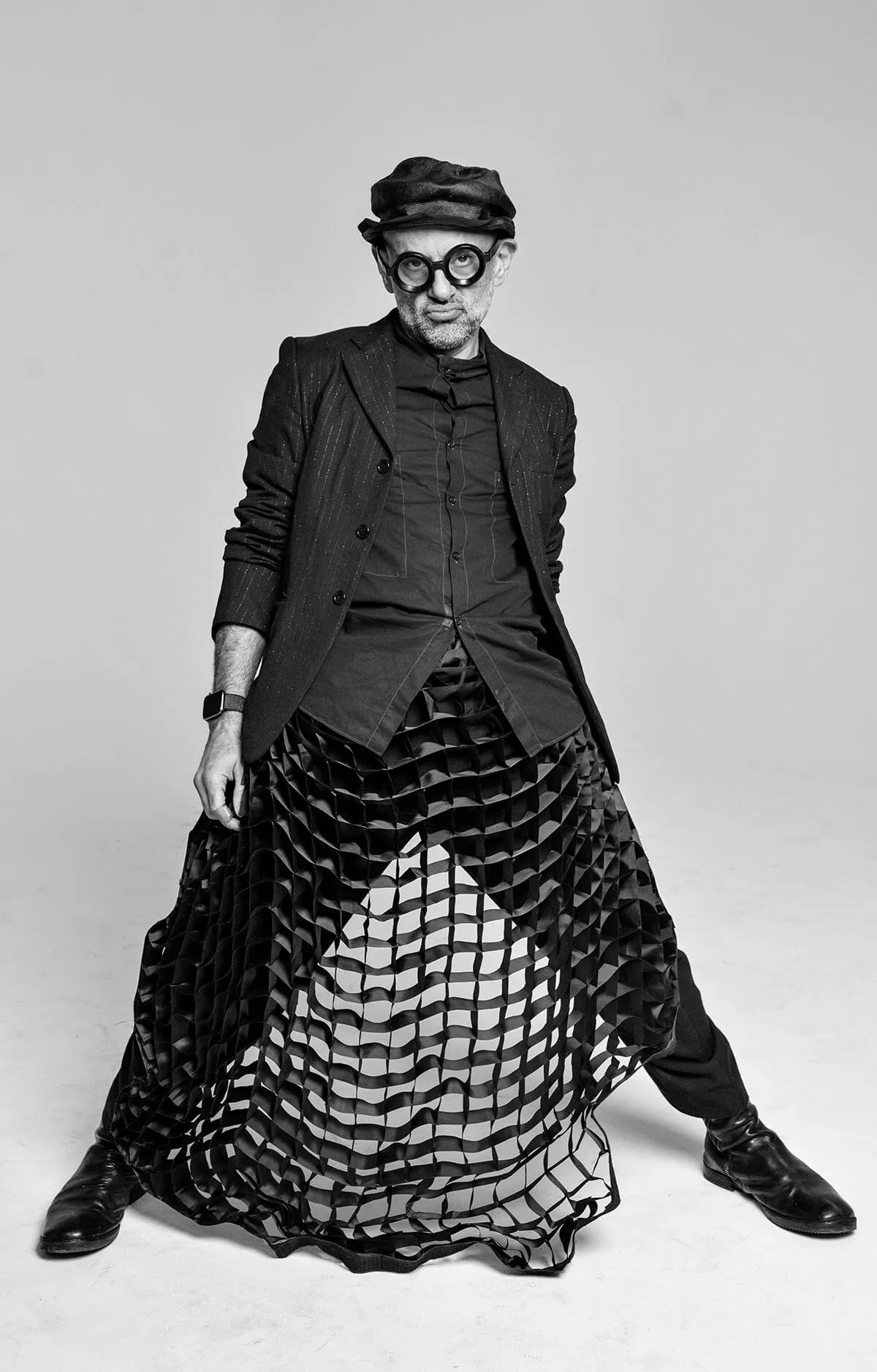 Black and white photo of a man wearing round glasses, a hat, a blazer, and a unique mesh skirt over pants and boots.