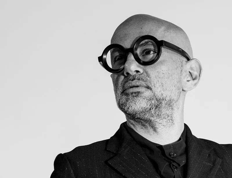 Black and white portrait of a bald man wearing round black glasses and a dark suit, looking slightly to the side.