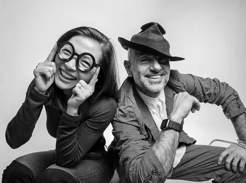 Black and white photo of a smiling woman wearing oversized round glasses and a man wearing a tilted hat, both posing playfully.