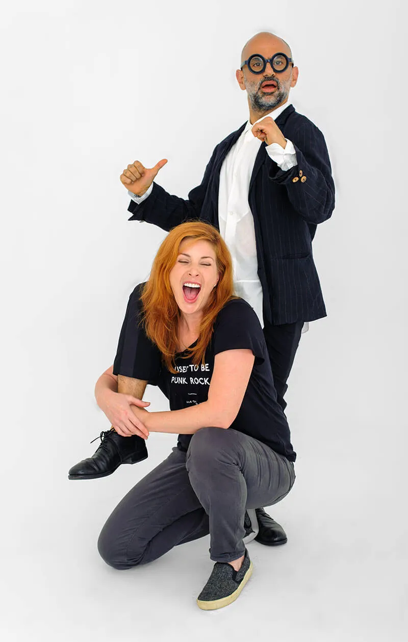A woman with red hair laughing energetically while hugging the leg of a man wearing a black pinstripe jacket and round glasses who looks surprised.