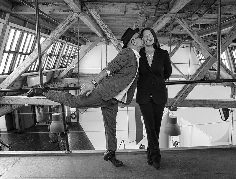 Man wearing a hat and patterned suit playfully kissing a smiling woman in a black pantsuit on the cheek in an industrial loft space.