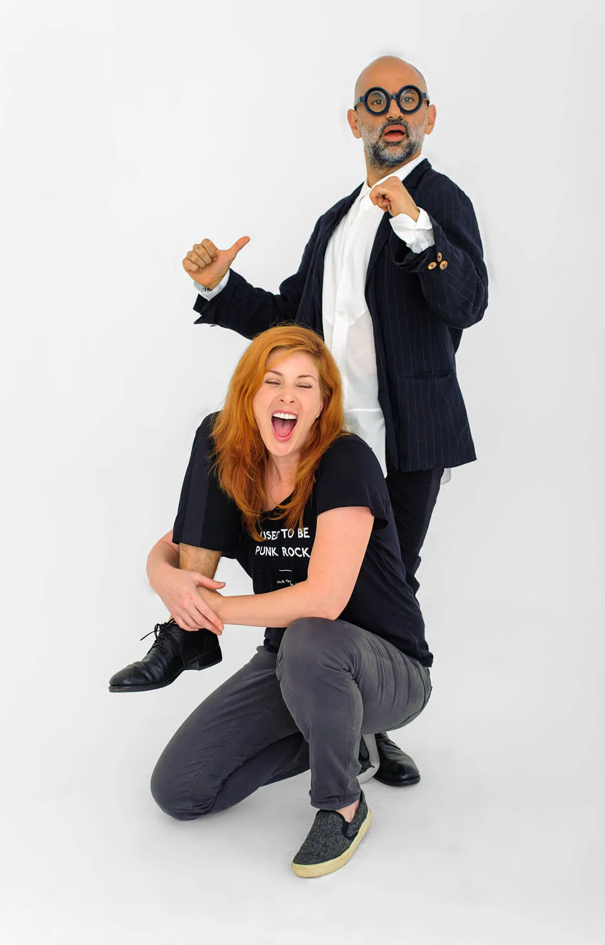 Woman with red hair laughing while hugging a man's leg who stands behind her wearing round glasses and a suit jacket.