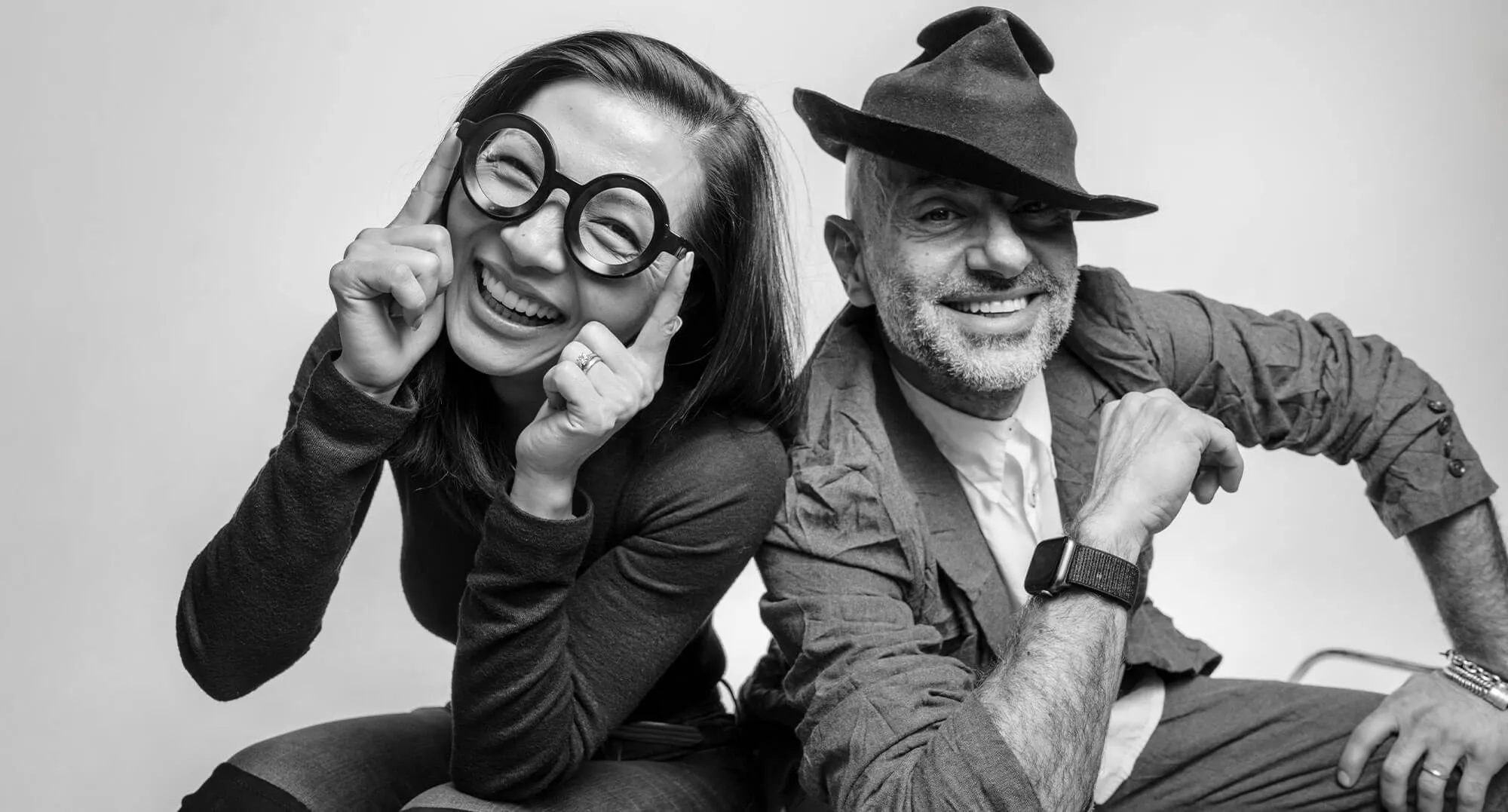 Smiling woman with round glasses and a cheerful man wearing a tilted hat posing together against a plain background.