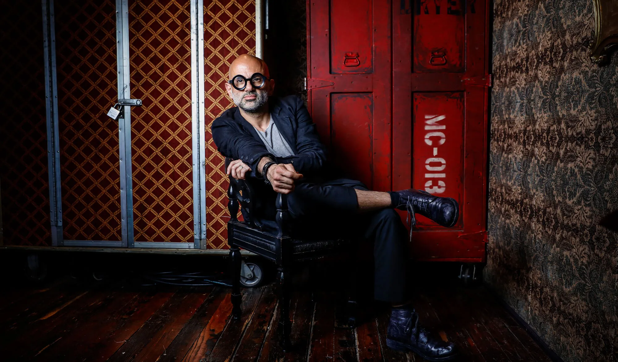 Man with round black glasses sitting cross-legged on a black chair against a red and patterned wall background.