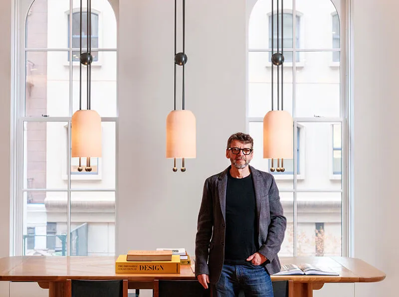 Man wearing glasses and a blazer standing in front of a wooden table with design books, with three pendant lights and large arched windows in the background.