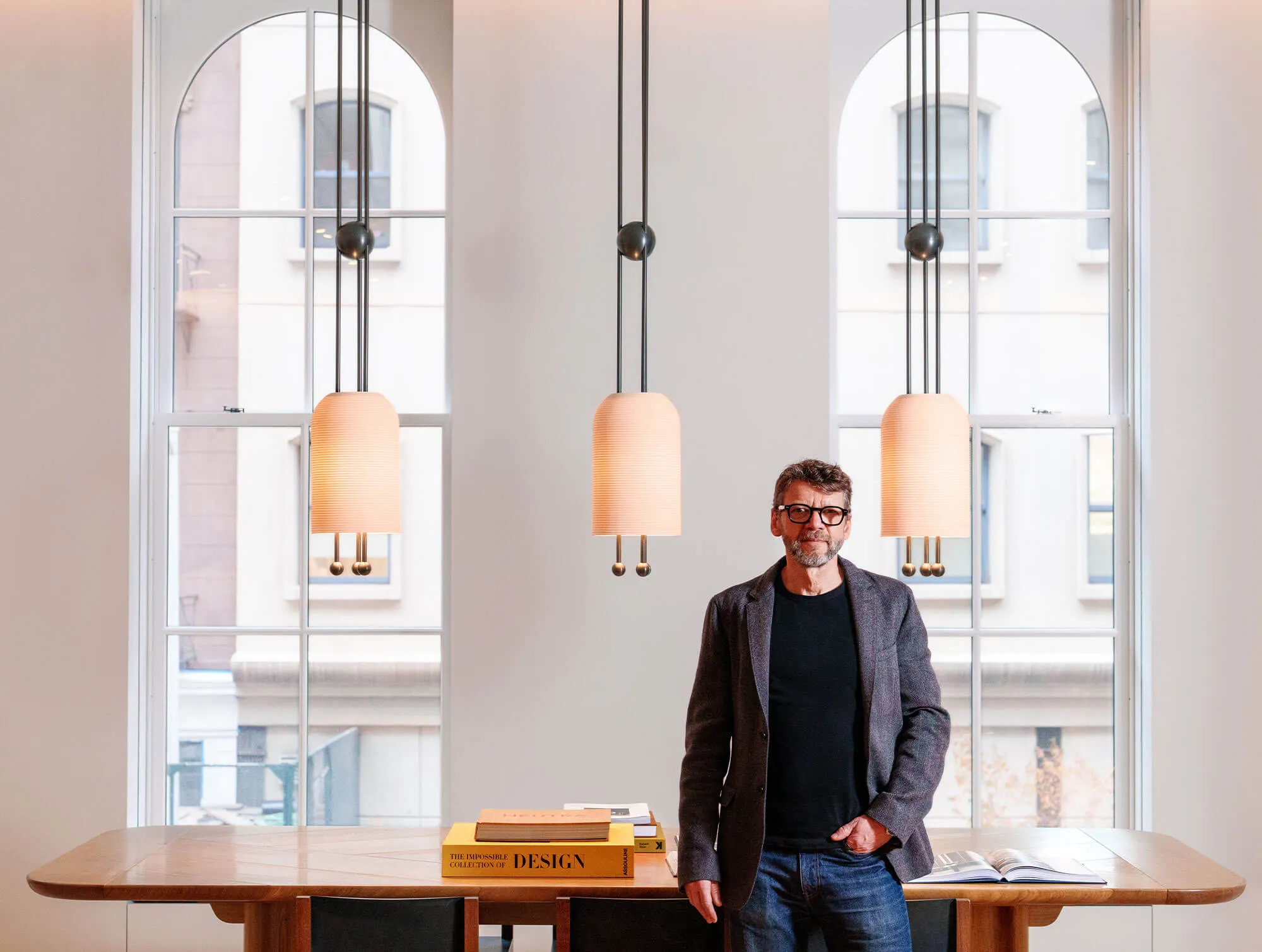Man with glasses, gray jacket, and jeans standing with hand in pocket in front of a wooden table with design books, large windows, and hanging pendant lights.