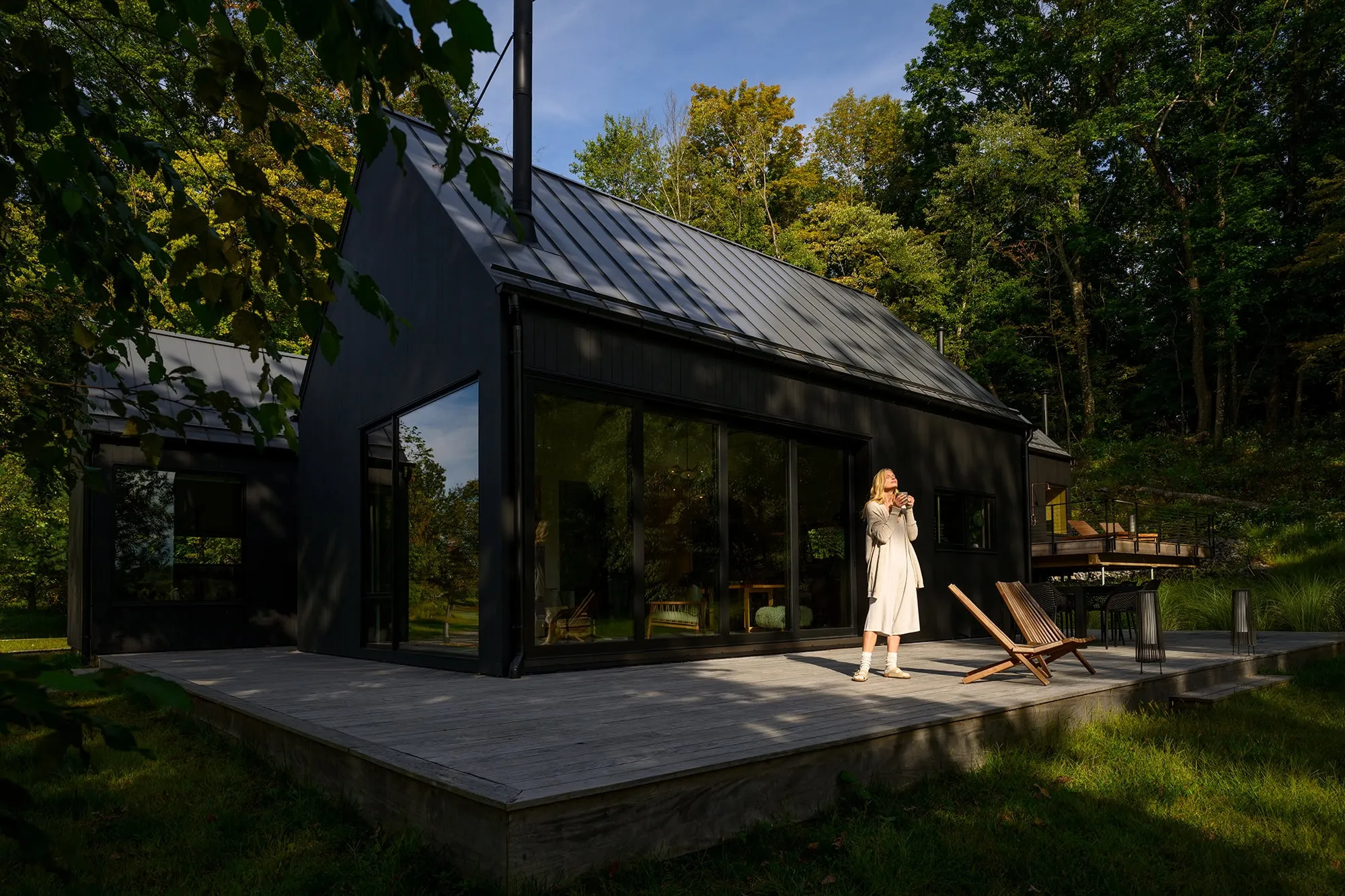 Modern black house with large glass windows on a wooden deck surrounded by green trees during daytime with a woman in a light dress standing on the deck holding a cup.