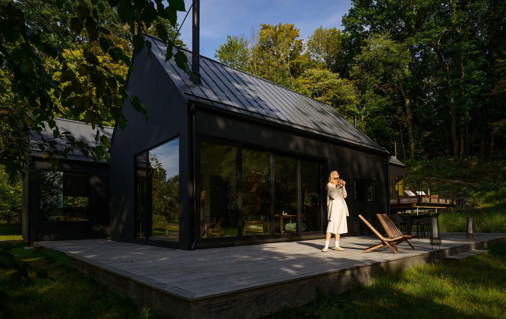 Modern black house with large glass windows on a wooden deck surrounded by green trees during daytime with a woman in a light dress standing on the deck holding a cup.