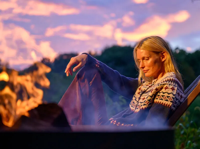 Woman in a patterned sweater sitting relaxed by an outdoor fire pit at sunset.