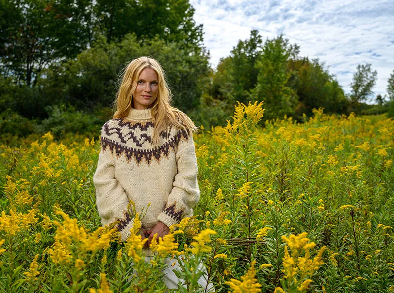 Woman wearing a cream-colored patterned sweater standing in a field of yellow wildflowers with trees in the background.