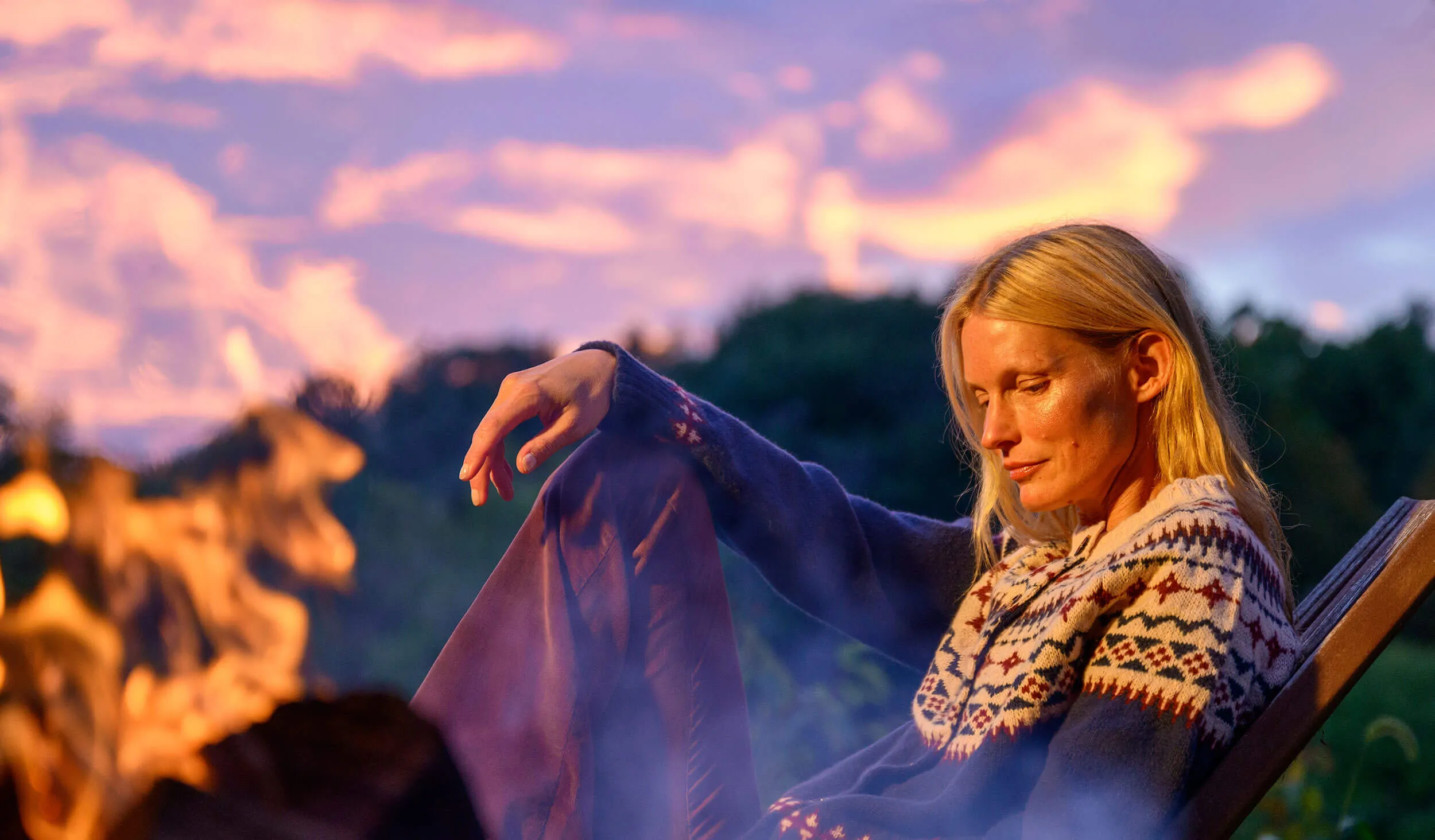 Blonde woman in patterned sweater relaxing by a campfire at sunset.