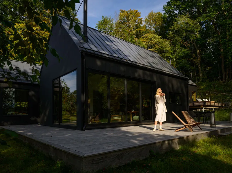 Woman in beige outfit standing on the wooden deck of a modern black house surrounded by green trees.