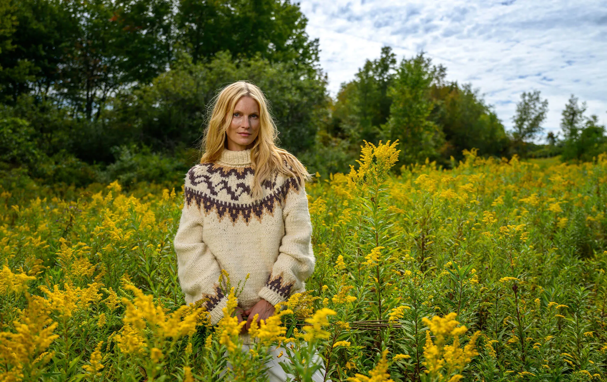 Woman with long blonde hair wearing a patterned wool sweater standing in a field of yellow wildflowers with trees and partly cloudy sky in the background.