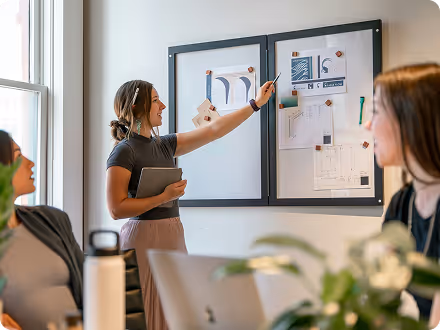 Woman presenting and pointing to notes on a whiteboard during a meeting with colleagues.