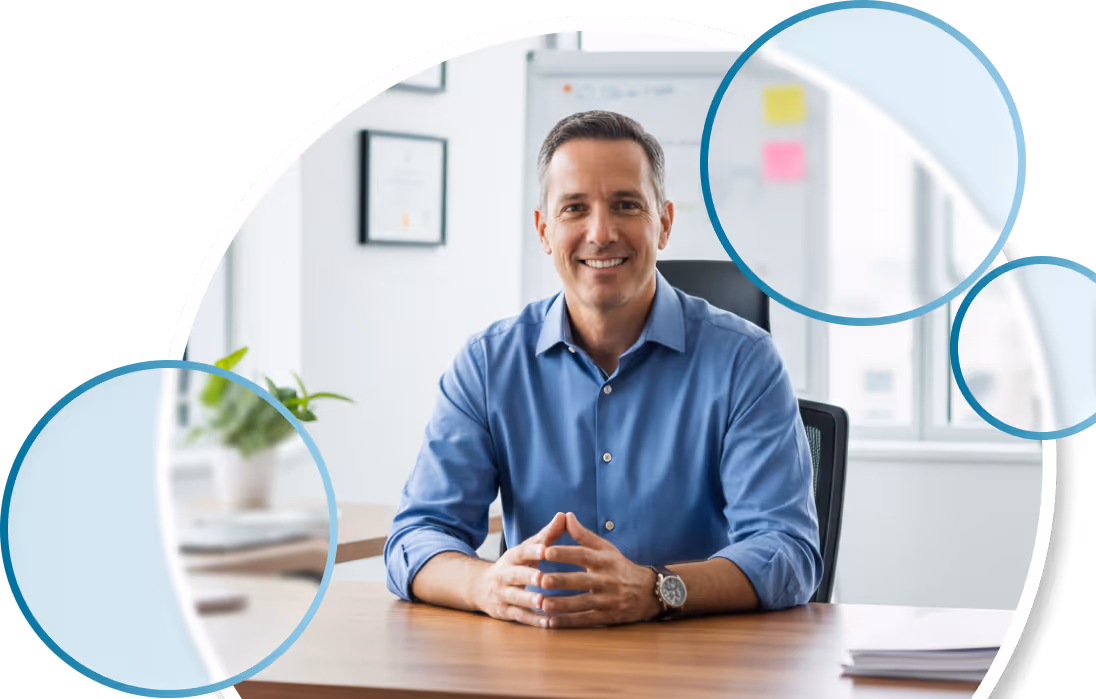 Smiling man in a blue shirt sitting at a wooden desk in a bright modern office.