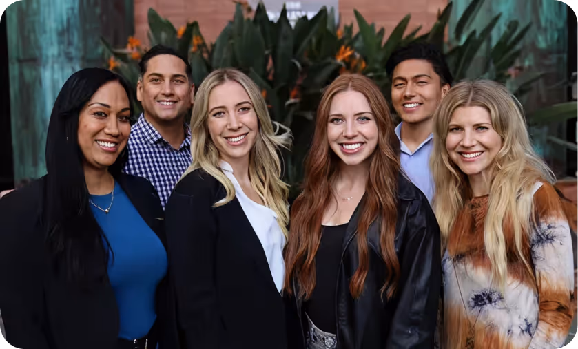Group of six smiling young adults posing outdoors with greenery in the background.