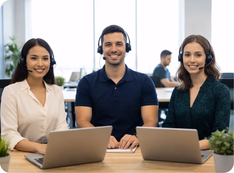 Three customer service representatives wearing headsets sitting at a desk with laptops, smiling at the camera in a modern office.