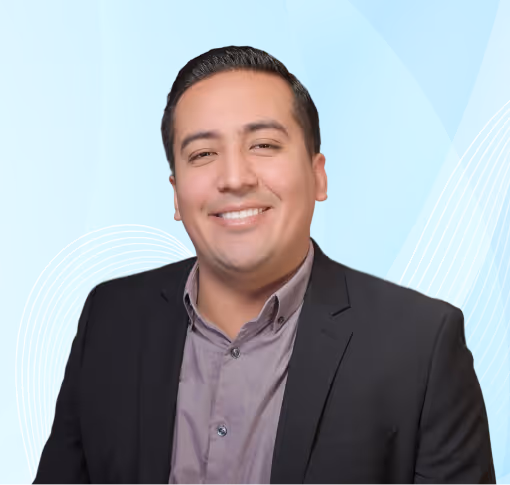 Smiling man with slicked-back dark hair wearing a black blazer and light purple shirt against a blue abstract background.