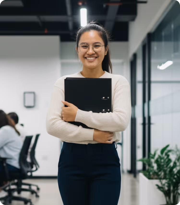 Smiling woman in glasses holding a black folder standing in a modern office.