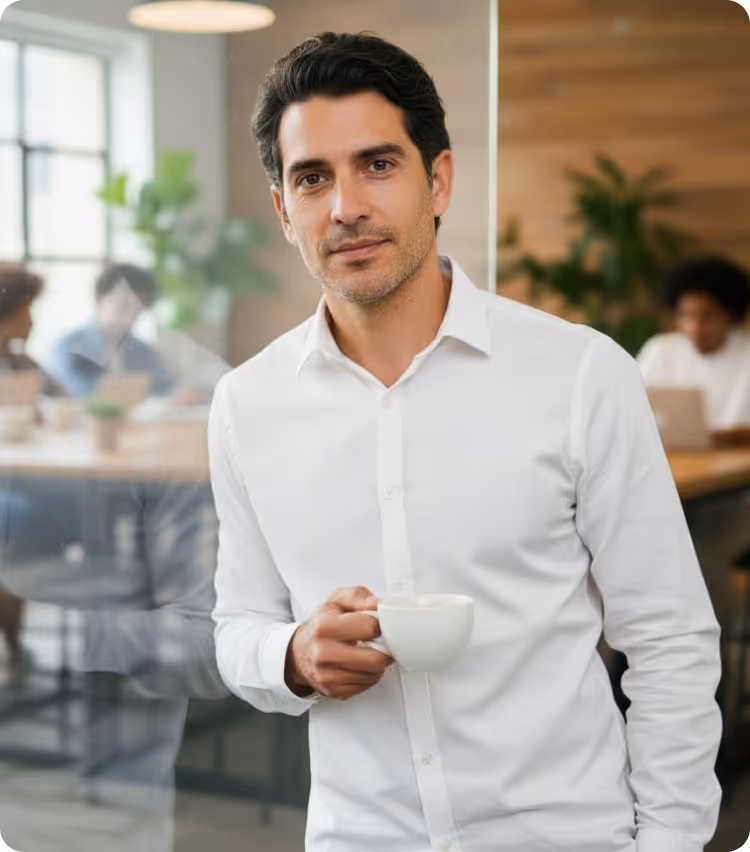 Man in a white shirt holding a cup, standing in an office with blurred coworkers in the background.