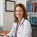 Female doctor with stethoscope around her neck smiling in an office with medical books and framed certificate on the wall.