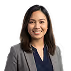 Smiling woman with shoulder-length dark hair wearing a navy blazer over a dark top, posing against a gray background.