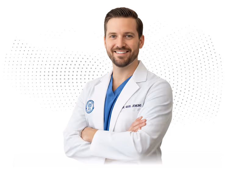 Smiling male dentist with arms crossed wearing a white coat and blue scrubs.