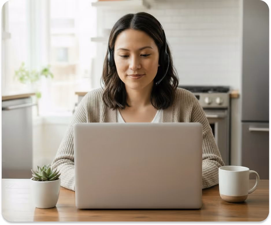 Woman wearing headset looking at laptop in kitchen with plant and coffee mug on table.
