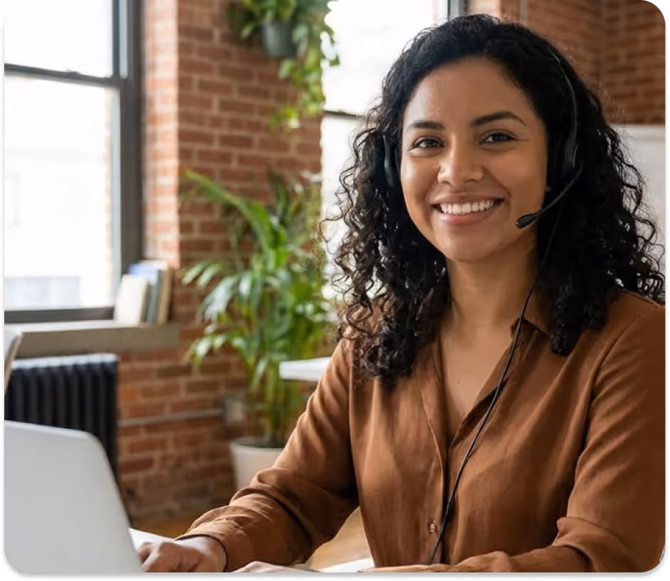 Smiling woman with curly hair wearing headset working on laptop in a bright office with plants.